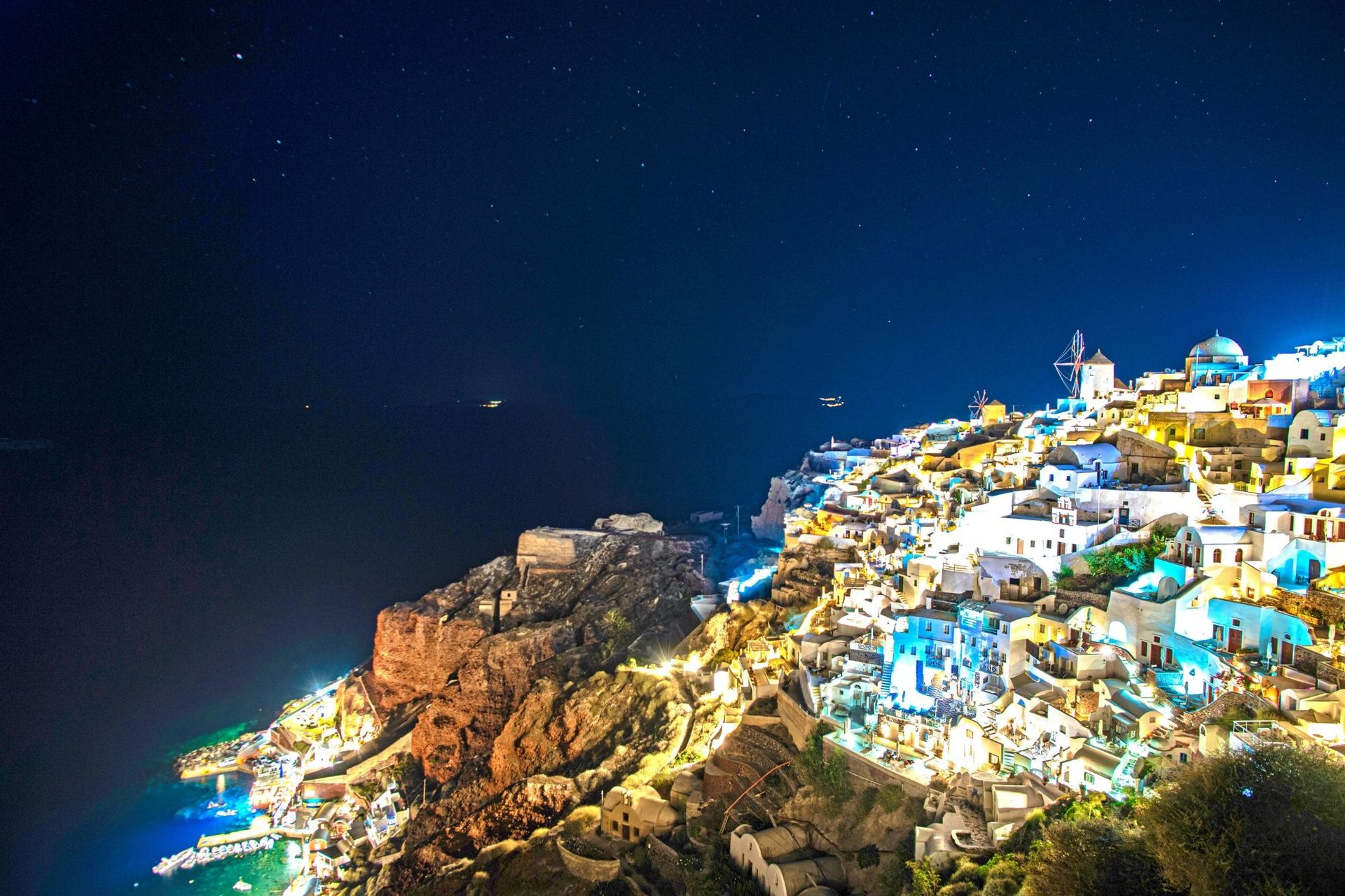 Oia, Santorini at night under a starry sky, the lit village on the cliff above the dark caldera sea